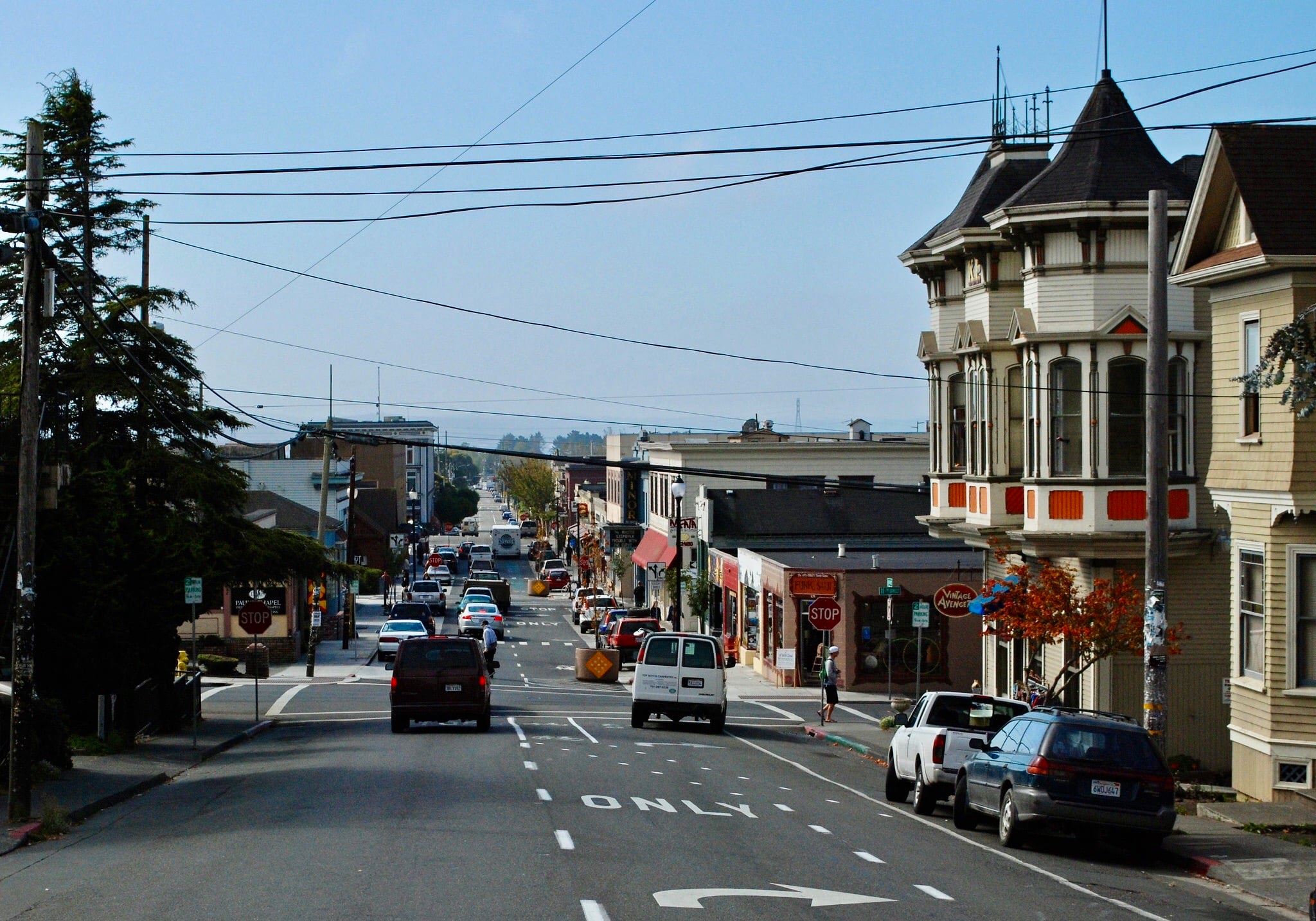A street in downtown Arcata with stores lining each side of the street. There are cars driving away and people crossing the street at a crosswalk