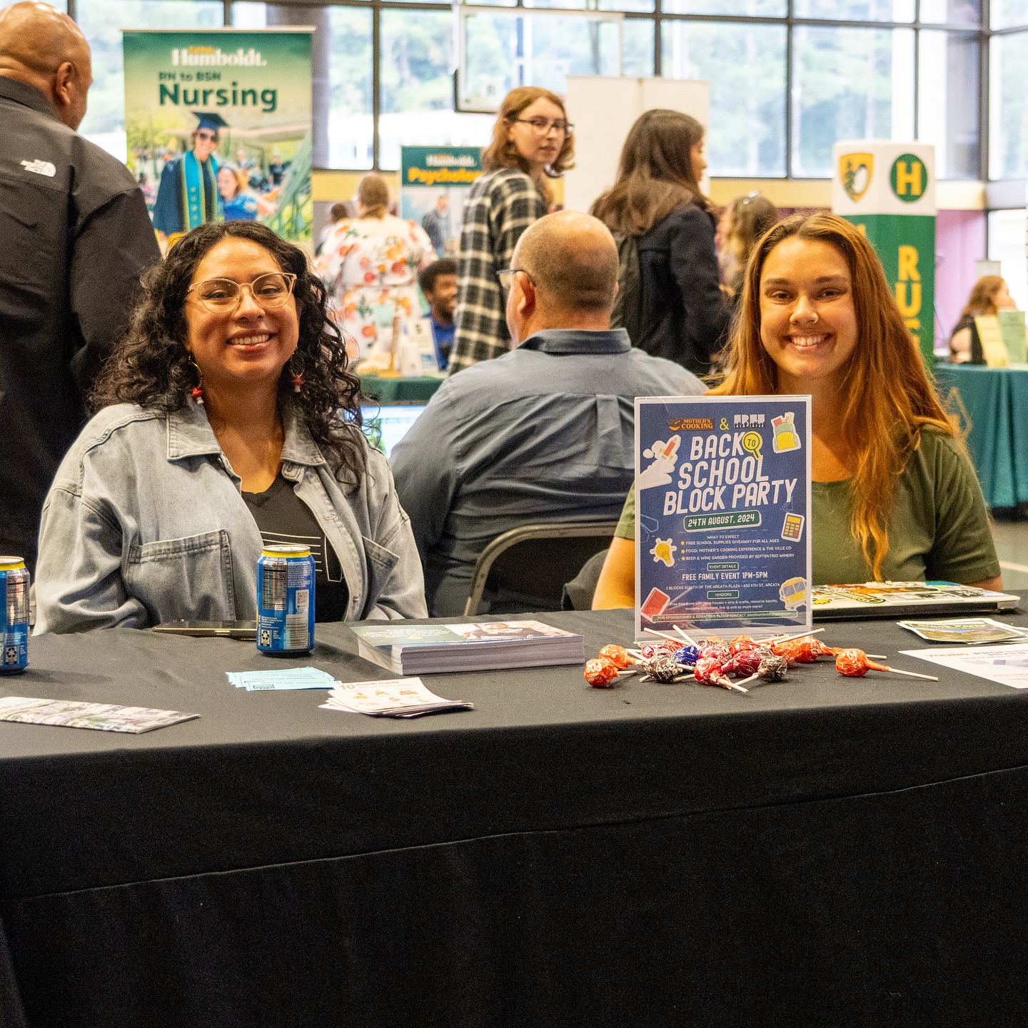 Two girls are sitting behind a table and smiling. On the table are lollipops and a sign that says "Back to School Block Party 24th August 2024, Free Family Event 1pm-5pm"