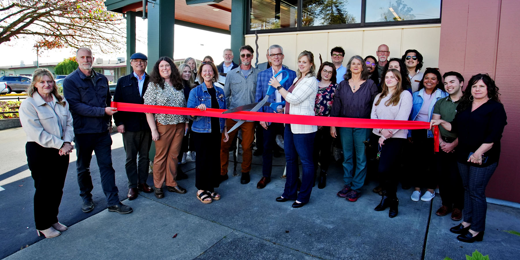 A group of 22 people are standing in front of a building, holding a red ribbon, which is being cut with giant scissors by the new business owner. The ribbon cutting ceremony is to celebrate and support new businesses as they open